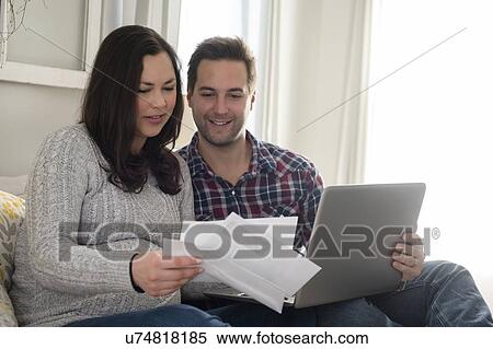 Stock Photography - Mid adult couple sitting on sofa and looking at letter, man holding laptop. Fotosearch