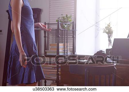 Stock Image - Mid adult woman, standing beside window, camera and laptop on table, mid section. Fotosearch