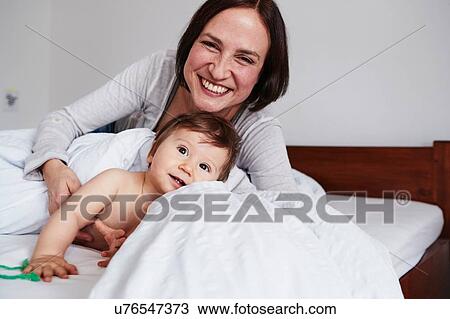 Stock Image - Mother and baby daughter relaxing on bed, smiling. Fotosearch
