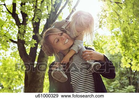 Stock Photo - Mother giving toddler daughter shoulder carry in sunlit park. Fotosearch