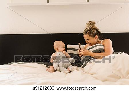 Stock Photograph - Mother photographing baby daughter sitting up in bed with soft toy. Fotosearch
