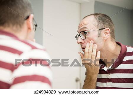 Stock Photo - Over shoulder view of mature man looking at his face in bathroom mirror. Fotosearch