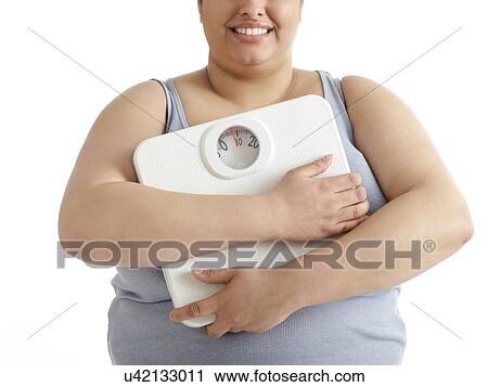 Stock Image - Overweight woman holding weighing scales, smiling.. Fotosearch