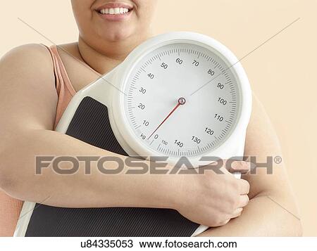 Stock Image - Overweight woman holding weighing scales, smiling.. Fotosearch
