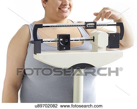 Stock Image - Overweight woman using weighing scales, smiling.. Fotosearch