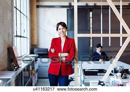Stock Photo - Portrait of businesswoman in office, arms folded, smiling, male colleague working in background. Fotosearch