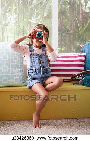 Stock Image - Portrait of girl on window seat photographing with toy camera. Fotosearch