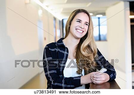Stock Image - Portrait of happy young businesswoman holding smartphone in office atrium. Fotosearch