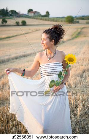 Stock Image - Portrait of mid adult woman, standing in field, holding sunflower. Fotosearch