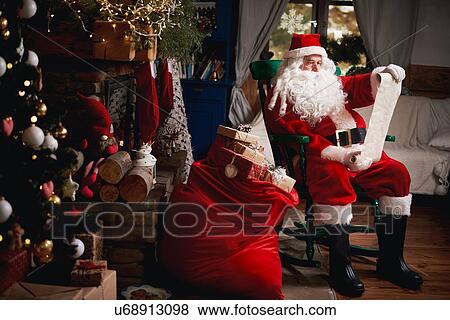 Stock Photo - Portrait of Santa Claus, sitting in chair with sack full of presents, looking at Christmas list. Fotosearch