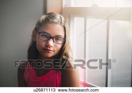 Stock Image - Portrait of young girl wearing glasses, standing beside window, pensive expression. Fotosearch