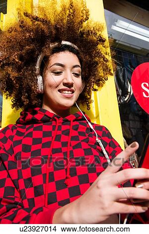Picture - Portrait of young woman outdoors, wearing headphones, holding smartphone. Fotosearch