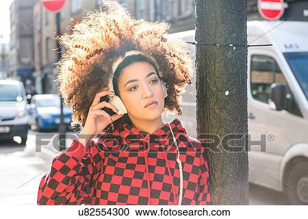 Stock Image - Portrait of young woman outdoors, wearing headphones, pensive expression. Fotosearch