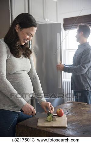 Stock Image - Pregnant woman cutting cucumber, man looking into refrigerator. Fotosearch