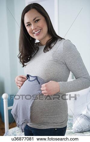 Stock Image - Pregnant woman holding infant bodysuit in bedroom. Fotosearch