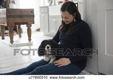 Stock Image - Pregnant woman sitting on floor and holding dog. Fotosearch
