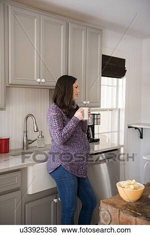 Stock Photo - Pregnant woman standing in kitchen and holding mug. Fotosearch