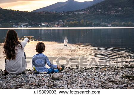 Stock Image - Rear view of boy and young woman throwing pebbles into river at dusk, Vercurago, Lombardy, Italy. Fotosearch