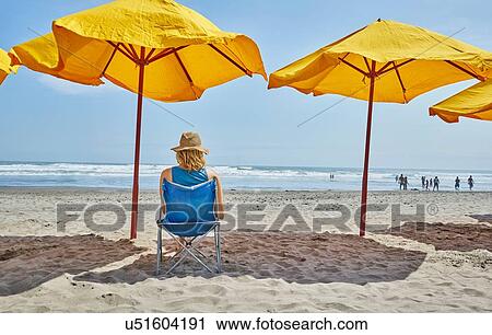 Stock Image - Rear view of female tourist sitting under beach umbrella, Camana, Arequipa, Peru. Fotosearch