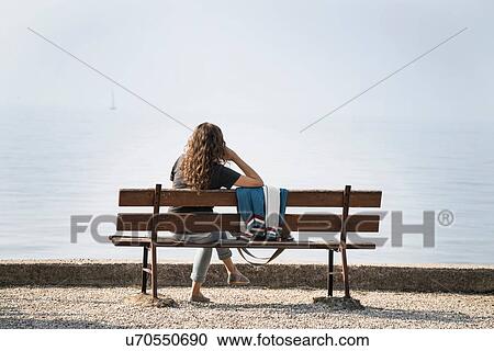 Stock Image - Rear view of woman sitting on bench looking at Lake Garda, Torri del Benaco, Veneto, Italy, Europe. Fotosearch