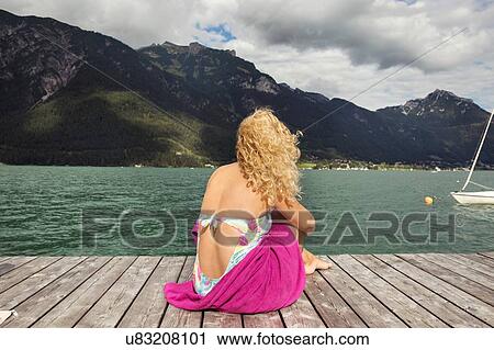 Stock Image - Rear view of woman sitting on pier looking away at view, Innsbruck, Tirol, Austria, Europe. Fotosearch