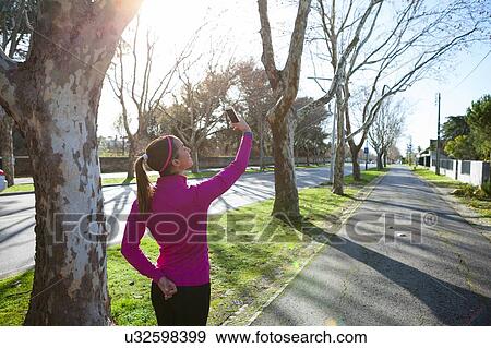 Rear view of woman taking selfie in tree lined street View Large Photo Image Stock Photo - Rear view of woman taking selfie in tree lined street. Fotosearch