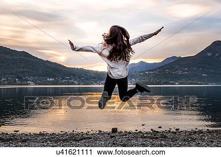 Stock Image - Rear view of young woman jumping mid air by river at dusk, Vercurago, Lombardy, Italy. Fotosearch