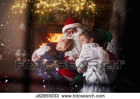 Stock Image - Santa Claus hugging young boy and girl, beside fireplace. Fotosearch