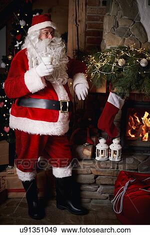 Stock Photo - Santa Claus standing beside fireplace, holding glass of milk. Fotosearch