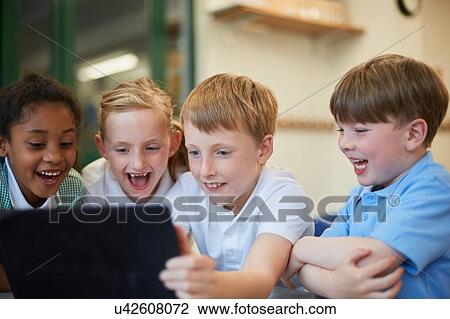 Stock Image - Schoolboys and girls laughing at digital tablet in classroom at primary school. Fotosearch