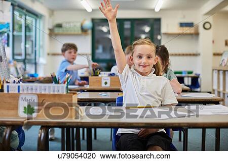 Stock Image - Schoolgirl with hand raised in classroom at primary school. Fotosearch