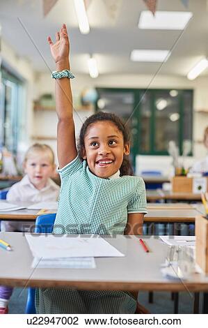 Stock Image - Schoolgirl with hand up in classroom at primary school. Fotosearch