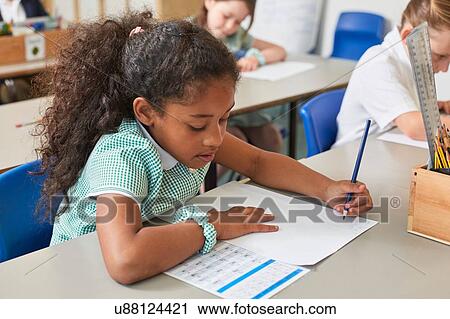 Stock Image - Schoolgirl writing at classroom desk in primary school lesson. Fotosearch
