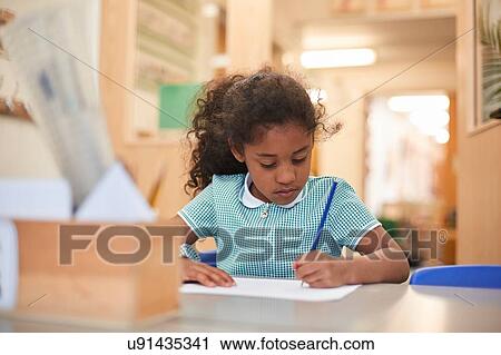 Stock Image - Schoolgirl writing at classroom desk in primary school. Fotosearch