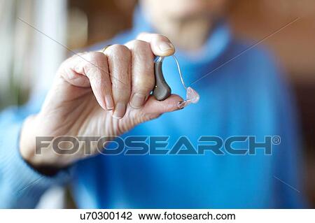Stock Image - Senior woman holding hearing aid, close-up. Fotosearch