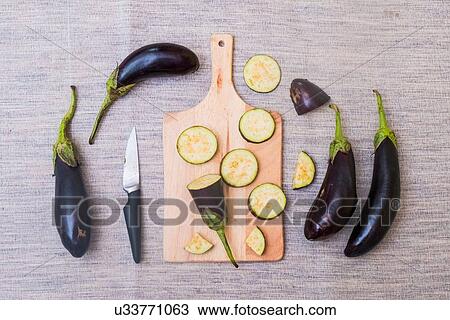 Stock Image - Slices of aubergine on chopping board, with knife and whole aubergines, overhead view. Fotosearch