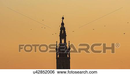 Stock Photograph - Spain, Andalusia, Seville, Bell tower of La Giralda against yellow sky with birds flying around. Fotosearch