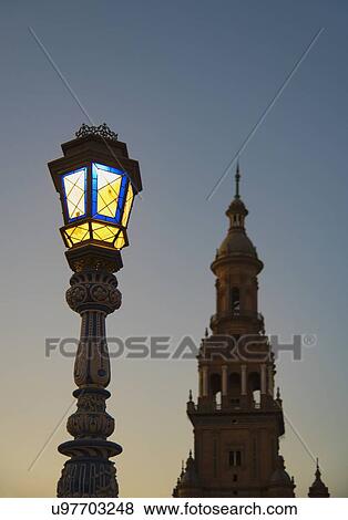 Stock Photo - Spain, Andalusia, Seville, Plaza de Espana, Illuminated old fashioned street lamp and tower at dawn. Fotosearch
