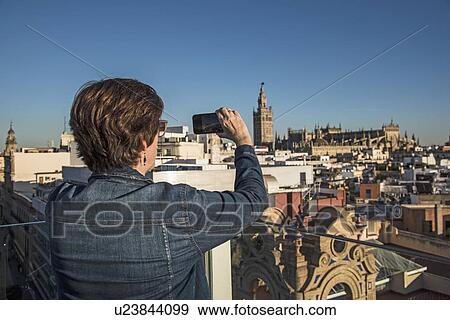 Stock Photo - Spain, Andalusia, Seville, Plaza Nueva, Woman taking photo of cityscape and la Giralda with smartphone. Fotosearch
