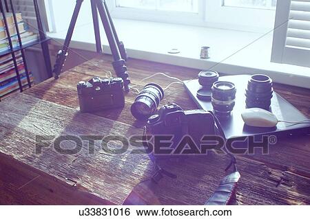 Stock Photograph - Still life of camera equipment and laptop on table in front of window. Fotosearch