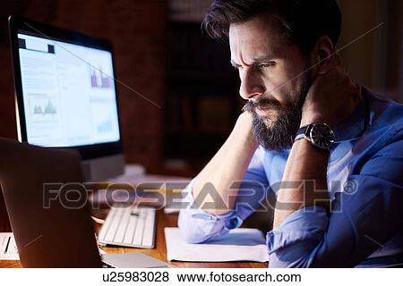 Stock Photo - Stressed young businessman looking at laptop on office desk at night. Fotosearch