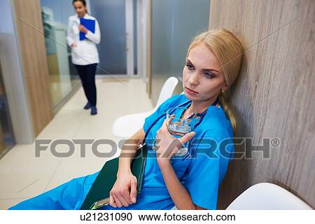 Stock Image - Surgeon sitting in hospital hallway, holding glass of water, pensive expression. Fotosearch