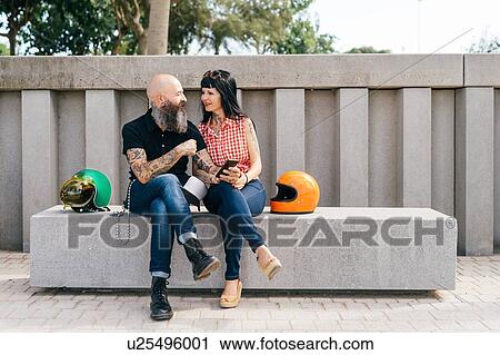 Stock Image - Tattooed mature hipster couple sitting on concrete bench. Fotosearch
