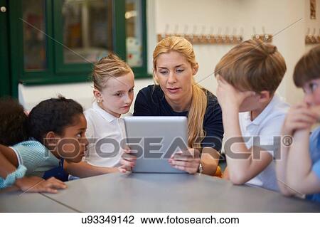 Stock Image - Teacher with schoolgirls and boys looking at digital tablet in classroom at primary school. Fotosearch