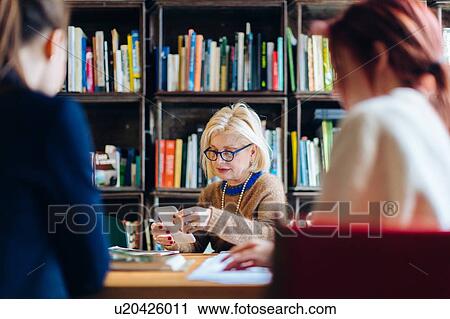 Stock Image - Three businesswomen sitting at table together, focus on woman using smartphone. Fotosearch