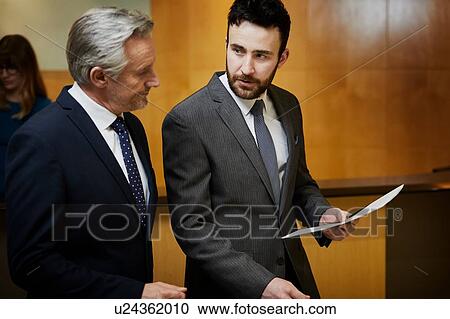 Stock Image - Two businessmen looking at paperwork in reception. Fotosearch