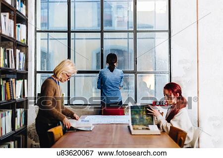 Stock Photo - Two businesswomen at table looking though books, another businesswoman looking out of window. Fotosearch