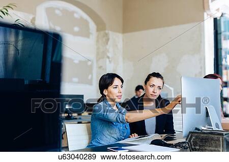 Stock Photo - Two businesswomen sitting at desk, looking at computer screen. Fotosearch