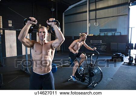 Stock Image - Two men exercising in gymnasium, using kettlebells and using air resistance exercise bike. Fotosearch