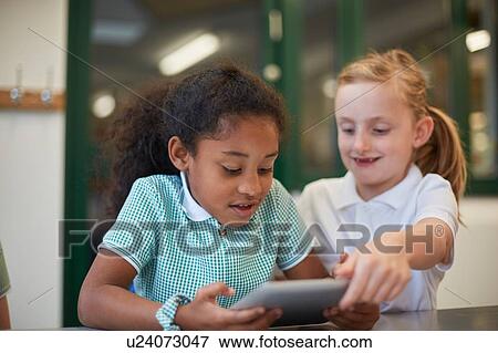 Stock Photo - Two schoolgirls looking at digital tablet in classroom at primary school. Fotosearch